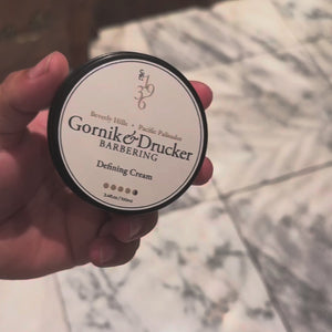 A barber opens the Defining Cream jar, works a scoop through damp hair and combs out a finished side part.